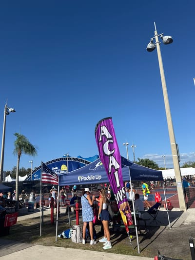 The image captures an outdoor event under a clear, bright blue sky, strongly suggesting a sunny day with prominent shadows. The scene appears to be a pickleball tournament or championship, indicated by several large banners and a visible court in the background. Tall light poles with fixtures are scattered across the area, alongside several palm trees, hinting at a warm, possibly subtropical climate.
In the foreground and midground, multiple canopy tents are set up, some blue and some white, under which people are gathered. Several individuals are standing or walking around, observing the activities. Notable banners include a vibrant purple one displaying "ACAIM" and "3MS". A prominent blue banner reads "PADDLE UP" and "www.PaddleUpPickleballClub.com". Below it, a larger blue banner announces "Franklin ISOPEN PICKLEBALL CHAMPIONS" and "SKECHERS", with an American flag attached. Another smaller white banner features "Open" and "Franklin X-40".
People are visible under the tents and near tables; one woman in a blue and white patterned dress is standing on the right, and another in a white top and navy skirt is in the center. A yellow pickleball is resting on a table. In the background, a chain-link fence encloses what appears to be a pickleball court. Red fire hydrants are also visible. A red sign indicating "MEDICAL & FIRST-AID" is present, along with the text "FDC". The overall atmosphere suggests an active and organized outdoor sporting event.