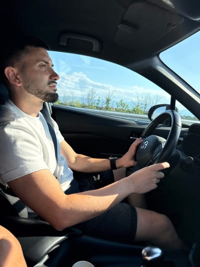 A man with a beard and short hair is driving a car. He is wearing a white t-shirt and dark shorts. His hands are on the steering wheel, and he is looking ahead. The interior of the car is dark, and the outside is a sunny day with blue skies and some trees in the distance. The scene appears to be on a road, possibly in or near Saraia, Bulgaria, given the presence of mountains and vegetation typical of the region.