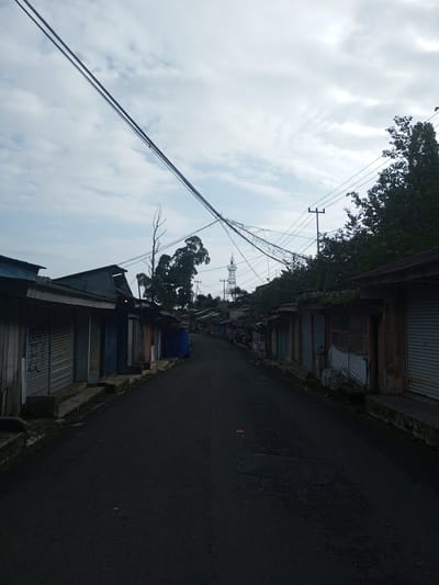 The image depicts a street in Mya Sit Kaing Ywar Thit, Myanmar. The street is paved and runs uphill. On either side of the street are buildings, primarily shops with corrugated metal shutters, most of which are closed. The buildings appear old and weathered. Overhead, numerous power lines crisscross the sky. Further up the street, a communication tower is visible. The sky is overcast. There are no people visible in the immediate foreground. The overall impression is of a quiet, possibly impoverished, and somewhat run-down location.