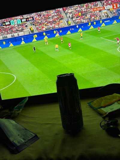 The image shows a television screen displaying a live broadcast of a soccer match between Australia (AUS) and Egypt (EGY), with the score at 0-1 in favor of Egypt. The game is taking place in a stadium with a large crowd in the background. On a surface in front of the television, there is a can of Monster Energy drink (zero sugar), two small packets of what appear to be snacks, and a set of keys. The scene is lit by the glow of the television screen, casting a greenish hue over the objects in the foreground. The overall setting suggests a casual viewing experience of the soccer match at a private residence. The location in the city of Gorni Dabnik, Bulgaria, is not explicitly depicted or suggested by any elements within the image.