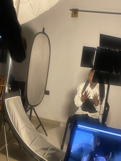 This is a medium shot of a person in a studio setting, likely in South Ropp, Nigeria, being photographed. The subject, dressed in a white collared shirt and black vest, is seated and looking towards the camera, with their hands held up, palms facing outward. The lighting setup includes a large softbox to the left, a reflector positioned behind the subject, and two large black panels that appear to be part of a lighting rig or backdrop. In the foreground, a laptop displays a preview of the photo shoot. The overall environment appears to be an indoor studio with a plain wall and a tiled floor.