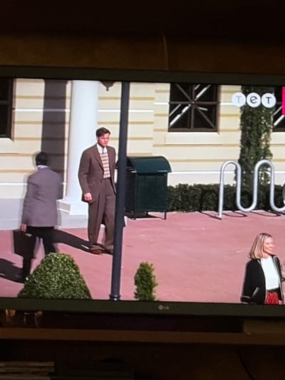 The image depicts a scene with several individuals and objects outside a building. A man in a brown suit and striped tie stands near a pillar. To his left, another man in a grey suit carries a briefcase and walks away from the camera. In the background, there is a dark green trash receptacle and a bicycle rack with several U-shaped supports. A woman with blonde hair, wearing a black jacket over a white shirt and a red skirt, is visible on the right side of the image. Green bushes are present in the foreground. The scene is set on a reddish-pink paved area in front of a building with light-colored walls and dark-paned windows. The overall environment appears to be an urban or public outdoor space. The image does not contain information that would place the scene specifically in Osolynka, Ukraine.