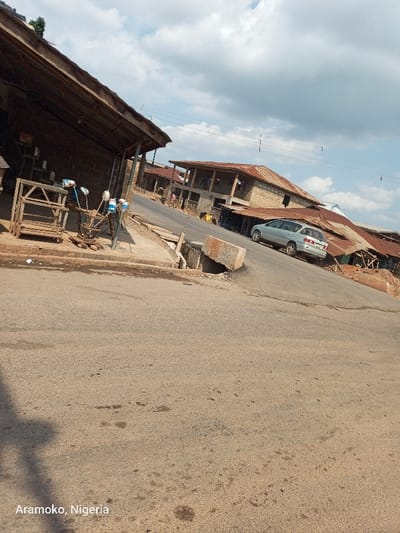 The image shows a street in Ilobu, Nigeria (7.837, 4.484), with a silver car parked on the right side of the street. There is a small, open-air market to the left of the street, with a few stalls and a wooden structure with a corrugated metal roof. The street is paved and dusty, with a drainage ditch on the left side. There are buildings in the background, including one with a corrugated metal roof. The sky is partly cloudy. There are no people visible.