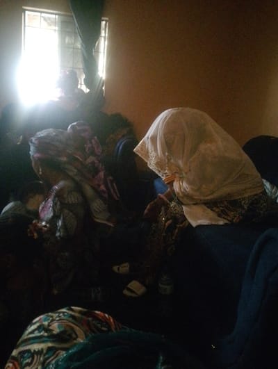 In a dimly lit room, individuals sit around a bed. One figure, draped in a white veil with gold trim, sits on the edge of the bed. Another individual, adorned in patterned fabric, sits to the left. Light streams through a window, casting shadows and obscuring details. Sandals and a water bottle sit on the floor. The room appears cluttered with various fabrics and garments.