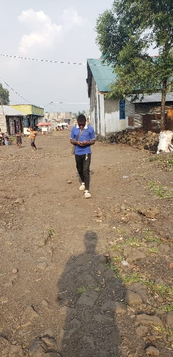 The image captures a daytime scene on an unpaved dirt road in Buzove, Democratic Republic of the Congo. In the foreground and center, a male individual, appearing to be an adult, walks forward, looking down and holding a smartphone with both hands. He is dressed in a blue short-sleeved collared shirt, dark trousers, and light-colored shoes. A dark strap, possibly from a bag, crosses his chest.

The ground consists of rough, dark brown earth mixed with rocks, debris, and sparse patches of dry grass. To the right of the path, a building with a light blue corrugated metal roof and white and unpainted concrete or stucco walls is visible. A small, blue-framed window is present on its facade. A large, green-leafed tree stands in front of this building, partially obscuring it. Near the base of the building, a pile of dark, irregular objects and a white sack are visible.

To the left, a series of lower-rise structures, likely commercial or residential, line the road, featuring various corrugated metal roofs and diverse wall materials. Further in the background, the dirt road continues, flanked by additional structures and faint figures, including what appear to be children playing. Utility lines are stretched across the upper portion of the frame against a hazy, partly cloudy sky. The distinct shadow of the photographer is cast on the ground directly in front of the viewer, extending towards the walking subject.