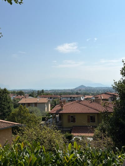 This image shows a view over a residential area in Soiano del Lago, Italy, with a hazy landscape in the background. In the foreground, lush green foliage is prominent, obscuring the lower parts of houses. Several houses with terracotta tile roofs are visible, some with yellow or beige exteriors and brown shutters. One house in the mid-ground has two satellite dishes on its roof. Beyond these houses, a town stretches out towards a body of water, likely Lake Garda, with mountainous terrain in the far distance, all softened by a light atmospheric haze. The sky is a pale blue with a few wispy clouds. The scene is captured during daylight, suggesting a clear but warm day. No people or animals are visible in the image.
