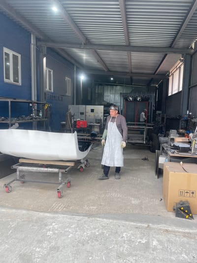 The image depicts a person, likely a mechanic or craftsman, wearing protective gear including safety glasses, ear protection, and an apron, standing in what appears to be a workshop or garage. The setting is industrial, characterized by corrugated metal walls and ceiling, concrete floors, and various tools and equipment.
In the foreground, a white, car bumper-like object is positioned on a metal stand with wheels, suggesting it is either being worked on or prepared for transport. In the background, there are shelves filled with items, machinery, and a large cardboard box with shipping symbols. A welding machine and gas cylinder are also visible, indicating that metal fabrication or repair work might be taking place.
The lighting suggests it is daytime, with artificial lights illuminating the interior. There is no visible indication of the weather outside. The overall atmosphere of the scene appears to be one of ongoing work and industrial activity. No specific emotions are evident, though the individual's posture suggests focus.
The image is oriented vertically. There is text on the cardboard box in the background, but it is not legible enough to be transcribed. The location context provided is Сокуры, Russia.