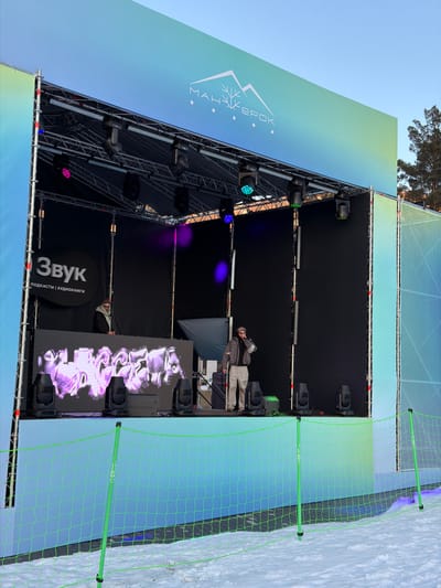 The image shows an outdoor stage setup in Озёрное, Russia. The stage is equipped with lighting fixtures, a large LED screen displaying graphics, and sound system elements. A person is standing on the stage, possibly adjusting equipment. The stage is surrounded by a net fence, and snow is visible on the ground, suggesting a winter event. The backdrop includes signage in Russian.