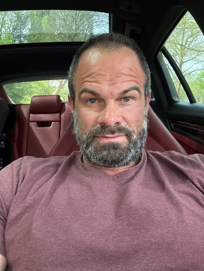 A man is sitting in the passenger seat of a car with a sunroof. He is wearing a purple t-shirt and has a graying beard. The car has red leather seats. Outside the window, trees are visible. The location cannot be determined to be in Diekirch, Luxembourg from the image alone.