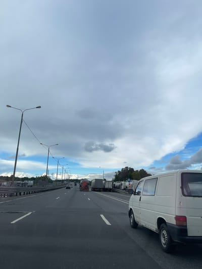 The image shows a highway in what appears to be Бавыкино, Russia, judging by the presence of Russian vehicles and roadside infrastructure. A white van occupies the right foreground, partially obscuring the view. The highway stretches into the distance with multiple lanes and is dotted with streetlights on both sides. Other vehicles, including trucks and cars, are visible on the highway, some seemingly parked on the shoulder. The sky is overcast with patches of blue interspersed with grey clouds. The road surface is asphalt, and the image is taken from a slightly elevated position, giving a wide view of the highway and surrounding environment.