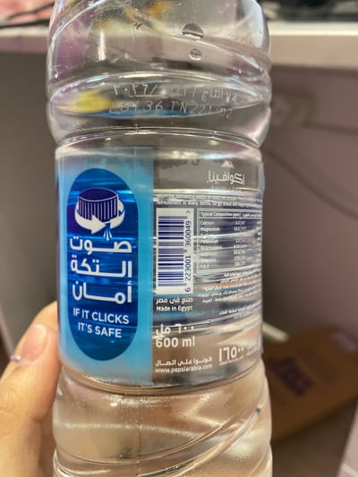 This media file is a close-up, vertical shot of a person's hand holding a clear plastic bottle of Aquafina drinking water. The bottle has a blue label with Arabic text and English text. The Arabic text translates to "If it clicks, it's safe." The English text reiterates this with "IF IT CLICKS IT'S SAFE." The bottle also displays nutritional information in both English and Arabic, detailing the typical composition of the water in parts per million (ppm), including Calcium, Magnesium, Sodium, Potassium, Bicarbonates, Sulphates, Chlorides, and Total Dissolved Solids (T.D.S.). There are also manufacturing details such as "Made in Egypt" and a website address, "www.pepsiarabia.com." The volume of the bottle is indicated as "600 ml."
The bottle also has a production code embossed on it, visible as "16136 TN22102" and a date "2026/05/11" (which appears to be a production date rather than an expiration date, given the year). The water inside the bottle appears clear with a few tiny bubbles. The background is blurred, suggesting an indoor setting, possibly a retail environment, with some brown cardboard boxes visible in the upper portion of the image. The lighting suggests it's daytime. The focus is on the product and its labeling.
