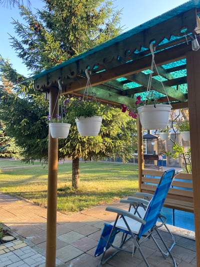 The image depicts a backyard scene with a wooden gazebo covered by a teal translucent roof. Several white hanging planters with purple and pink flowers are suspended from the gazebo's structure. In the foreground, two blue and white striped folding chairs are positioned on a brick-paved area. Behind the chairs, a wooden bench is visible. The background features lush green trees, including pine trees with visible cones, and a grassy lawn. The sun is casting shadows, indicating it is either early morning or late afternoon. There are no people visible in the immediate vicinity of the gazebo or chairs. The environment suggests a residential backyard, possibly in a suburban or rural setting, given the mature trees. The location is identified as Volzhsk, Russia, which is a city in Mari El Republic known for its industrial areas and proximity to the Volga River.
