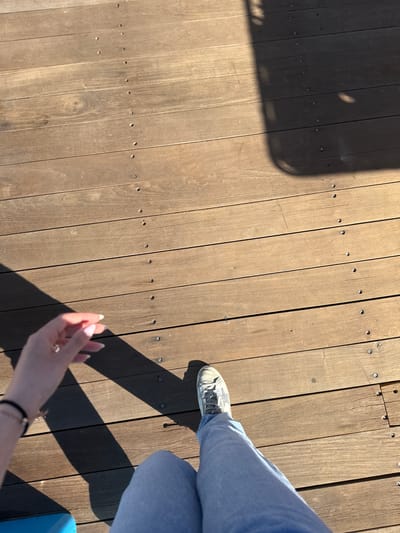 A first-person, high-angle shot captures a wooden deck surface in daylight. The deck is made of horizontal wooden planks secured with a line of visible screw or bolt heads along each plank. A shadow is cast on the left side of the image. Part of a person is visible in the right side; clad in light blue denim pants and a light-colored shoe. A hand is also visible with light-colored nails. The environment suggests an outdoor setting, potentially a pier or boardwalk. The location in La Paz, Mexico cannot be verified from the visual elements alone.