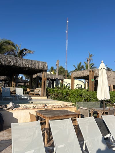 This image depicts a sunny outdoor area, likely part of a resort or beachfront establishment, under a clear blue sky in Mazatlán, Mexico. The bright sunlight suggests it's midday or early afternoon, casting distinct shadows across the scene.
In the foreground and extending into the mid-ground on the right, several empty lounge chairs with light grey fabric are arranged on a paved patio or deck. To the left of these chairs, a walkway leads to a series of shaded structures. These structures feature traditional thatched roofs, known as palapas, supported by robust wooden posts, providing a tropical ambiance. Underneath the palapas, wooden tables and chairs are arranged for outdoor dining or relaxation. A large, closed white umbrella stands near one of the shaded areas.
Lush green foliage, including manicured bushes and several tall palm trees, is interspersed throughout the scene, contributing to the resort-like setting. In the background, a light-colored building with distinctive arched windows is visible, likely part of the main complex. A small green sign with a white arrow pointing to the right is affixed to one of the wooden posts within the shaded area. On the far left, a tall, slender structure, possibly a communication tower or a distant crane, extends high into the clear sky. The area appears quiet and ready for guests, with no people visible in the frame.