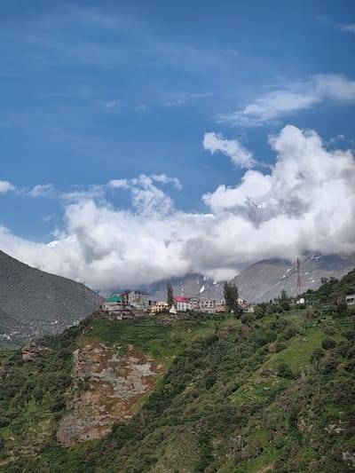 The image depicts a high-altitude village nestled on a mountainside in Kardang, India. The scene is dominated by a lush green slope in the foreground, interspersed with trees and rocky outcrops. A cluster of buildings, characterized by multi-story structures with colorful roofs, is visible on the crest of the hill, forming the village. Behind the village, a range of majestic mountains rises, with their peaks partially obscured by dramatic, white clouds. Snow can be seen dusting some of the higher slopes. The sky above is a vibrant blue, with scattered white clouds creating a dynamic atmosphere. A tall telecommunications tower stands prominently on the right side of the frame, suggesting modern infrastructure within this remote setting. The overall impression is one of a serene, natural landscape with a human settlement harmoniously integrated into its surroundings.