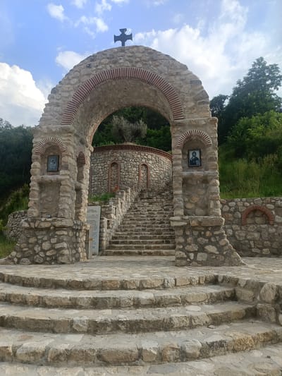 This image depicts an exterior view of an arched stone entranceway leading to a circular stone building with a staircase. The entrance is adorned with brickwork and has a dark cross at its apex. Flanking the archway are stone pillars, each featuring a framed icon of a religious figure.

The scene is set outdoors, with a lush green hillside and trees forming the backdrop. The sky above is partly cloudy, suggesting daytime. The architecture, characterized by rough-hewn stones and decorative brick arches, gives the impression of an ancient or historic site, possibly a monastery or religious sanctuary, consistent with the location context of Melnik, Bulgaria.

A sign with visible text is placed to the right of the staircase. While the text is partially obscured and small, it appears to be in Cyrillic script, further supporting the Bulgarian location. The steps leading up to the circular structure are also made of stone and appear weathered, contributing to the rustic and historical ambiance. The overall mood conveyed is one of serenity and reverence, with a hint of historical significance. No people are visible in the frame, and the activity appears to be stationary, focused on the architecture and its surroundings.