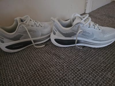 This image captures an indoor scene in Shotton, United Kingdom, displaying two white Nike athletic shoes resting on a grey, low-pile carpet. The shoes are oriented with their toes facing right, positioned against a plain, light-colored wall. A segment of a white doorframe is visible on the far right side of the background.
The shoe on the left exhibits minor signs of wear, featuring a white mesh upper with dark grey horizontal stripe accents on its sides. A grey Nike "swoosh" logo is present on its lateral side, and "ZoomX" is visible in white text on the dark grey or black sole insert. The laces are white and partially tied.
The shoe on the right appears cleaner, also featuring a white mesh upper with dark grey horizontal stripe accents and a grey Nike "swoosh" logo on its lateral side. Its midsole is white with a prominent black, wavy-patterned insert. The white laces are untied and draped across the top of the shoe.
