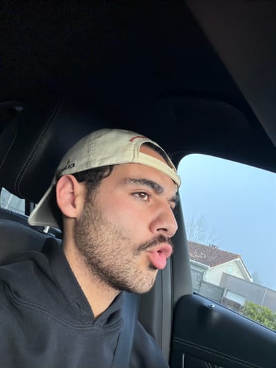 The image shows a young adult male inside the passenger compartment of a vehicle. He possesses dark hair, brown eyes, and facial stubble, including a mustache and beard. He is wearing a beige baseball cap backward, which features "ROLEX" lettering on the left side and a red logo on the rear strap. His attire includes a dark-colored hooded sweatshirt, and a dark seatbelt is fastened across his left shoulder and chest. The man is looking towards the right, with his mouth open in a puckered expression, and his tongue slightly extended.

The vehicle's interior is dark, with a black headliner and a black leather seat with visible stitching behind the man's head. The right side of the frame reveals a black door panel and window frame.

Through the vehicle's window, an external environment appears hazy or foggy. A residential structure with a distinctive orange-brown tiled roof and light-colored walls is partially visible in the background. Bare tree branches are also discernible. While the location is specified as Mont-de-Marsan, France, the specific street or detailed address within the city is not identifiable from the image. The scene depicts a suburban residential area.