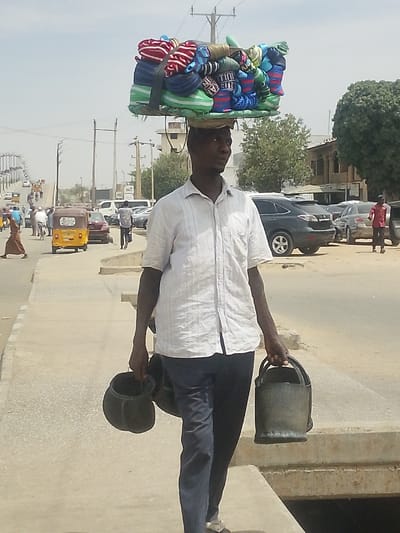 The scene depicts a daytime street in Gyadi Gyadi Arewa, Nigeria, where an adult male walks on a paved sidewalk. He wears a light-colored, short-sleeved collared shirt with faint vertical stripes and dark trousers. A large, rectangular stack of numerous folded, colorful knitted caps or hats, secured with green straps, is balanced on a round pad on his head. He holds a dark, cylindrical container, resembling a bucket, in each hand. In the immediate background, a multi-lane paved road displays moderate vehicle traffic, including various cars and a yellow three-wheeled auto-rickshaw. Several other pedestrians are visible on or near the road. Utility poles with overhead power lines line the street, while various urban buildings and scattered green trees populate the mid-ground and distant background. A concrete drainage channel runs parallel to the sidewalk in the foreground.