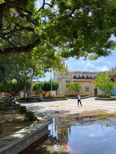 The image captures a wide shot of a town square in San José de Barlovento, Venezuela. A prominent yellow church with a clock tower dominates the background, its reflection visible in a large puddle on the tiled ground. Lush green trees, including palm trees, provide shade throughout the square. A young person, dressed in a white t-shirt and dark pants, walks across the square. Several other individuals are seated on benches around the perimeter of the square, some appearing to be engaged in conversation or simply relaxing. The sky is clear with scattered clouds, suggesting a pleasant day. The paving stones of the square are interspersed with red tiles, creating a decorative pattern. The overall scene depicts a tranquil public space within the town.