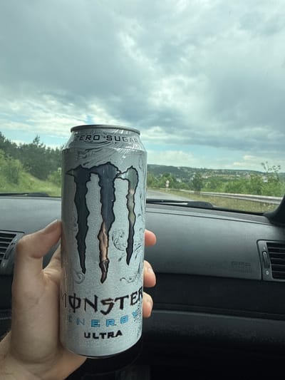 A hand holds a can of Monster Ultra Zero Sugar energy drink inside a car. The car's dashboard is visible, and the exterior landscape of trees and sky suggests a road trip. The location cannot be determined as Novi Pazar is a city in both Bulgaria and Serbia.