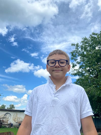 A boy wearing glasses and a white polo shirt is photographed outdoors against a backdrop of a bright blue sky with scattered white clouds. To his right, a lush green tree with visible branches and leaves occupies the frame. In the lower left quadrant, part of a white wall with a crude drawing of a face and some structures is visible. The overall scene suggests a casual, perhaps rural, setting.