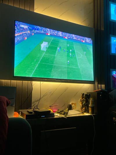 The scene portrays an indoor entertainment area within Oko Town, Nigeria, dimly illuminated by a warm yellow ambient light strip situated beneath a large, wall-mounted flat-screen television. The television displays a football video game or broadcast showing a match between PSG and BAR, with the score tied at 1-1 at the 23:02 mark. The screen features a green football pitch, a goal, multiple player figures in blue and white kits, and a stadium backdrop. Beneath the television, on a dark console, are several electronic devices, including a "SPROIC" unit, a remote control, and associated wiring. Decorative elements on the console include an orange globular object, a golden ornate statue depicting a figure with an instrument, two bottles (one yellow, one white), and a dark crumpled fabric. The wall directly behind the console features light-colored, possibly marble-patterned panels, while a darker, vertically paneled section is visible to the right, adjacent to multiple square wall frames containing blue-tinted or lit objects. A portion of a teal-colored upholstered item, likely a chair or sofa, is visible in the left foreground. No individuals are present in the observable frame.