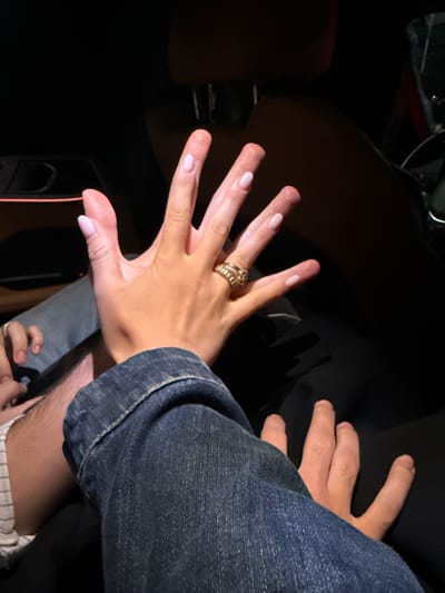 The image depicts two hands, one with a gold ring on the ring finger, against a dark background with hints of a car interior. The hand with the ring is outstretched. The other hand, partly visible, is partially obscured by a denim sleeve. The location is unknown.