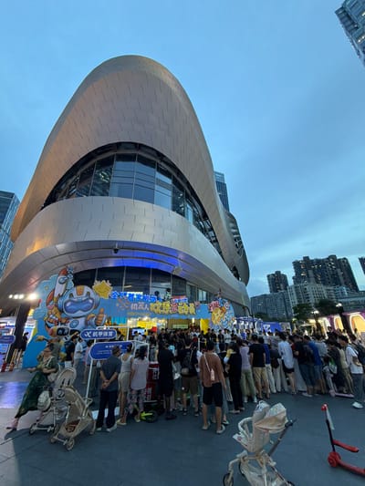 A group of people are gathered outside a modern building with a curved, silver facade. The building's glass windows reflect the overcast sky and surrounding cityscape, which includes tall apartment buildings in the background. The gathering appears to be an outdoor event, possibly a festival or exhibition, indicated by the colorful banners and displays set up along the building's exterior.
One banner features cartoon characters, including an astronaut, a rocket, and a smiling planet, suggesting a theme related to space or science. Text on the banner is in Chinese, with some English text also visible, such as "ROBOT FUN FEST". The overall atmosphere seems to be lively, with many people standing and looking towards the displays.
Several strollers are present, indicating that families with young children are attending the event. A red scooter is parked nearby. The lighting suggests it is late afternoon or early evening, and the sky is cloudy, creating a diffused light. The location is identified as Longgang District, China. The event appears to be family-friendly and engaging.