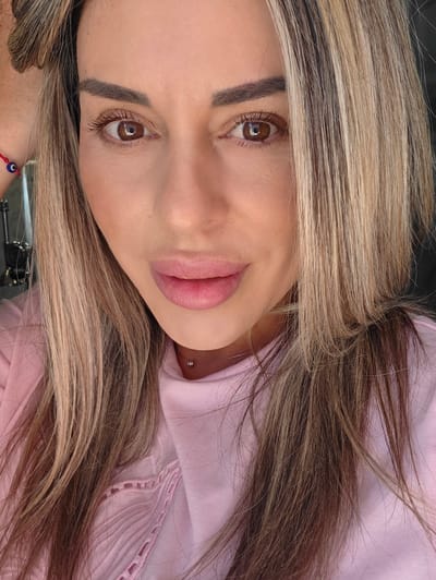 A close-up, head-and-shoulders portrait of a woman with blonde and brown highlights in her hair. She has brown eyes, dark eyebrows, and is wearing pink lipstick. She has a silver stud piercing on her neck. She is wearing a pink shirt. The background is dark and out of focus. No specific location in Carpi, Italy is discernible.