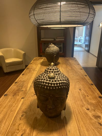 This image depicts an interior residential space, likely a dining or living area, located in Staudernheim, Germany. The scene is lit by warm, ambient overhead light. A long, rectangular wooden table with a prominent grain and dark circular inlays occupies the foreground and midground. Two decorative Buddha head statues are placed along the table's centerline. The statue in the foreground is a dark, textured bronze or antique gold color, featuring closed eyes, tightly curled hair-like protuberances, and a prominent, golden ushnisha. A second, smaller statue of similar design is positioned further down the table.

A dark, elliptical pendant light fixture, constructed from interwoven material, hangs centrally from the ceiling above the table. To the left of the table, an off-white upholstered armchair is positioned against a plain, light-colored wall. In the central background, a dark wooden bookshelf contains several books and items, including one with "GREEN" visible on its spine. To the right, an open doorway reveals a brighter, adjacent room or hallway containing light-colored furniture and a decorative, illuminated metal wall fixture. No people are present in the scene.