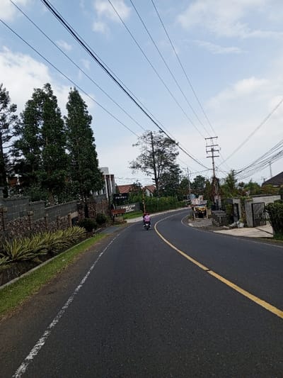 The image depicts a daytime scene in Ciloto, Indonesia, showing a two-lane asphalt road curving to the right. A single individual, wearing a pink top and a helmet, is operating a black scooter, traveling away from the viewer along the right side of the road. The road features a solid yellow center line and a solid white shoulder line on the right.
To the left of the road, a strip of decorative yellow-green plants is bordered by a dark stone wall. Behind the wall are several tall, slender evergreen trees. Further back, a brown sign with white text reads "GRANDMISION PARK & RESORT." A building with a dark roof is partially visible behind the trees and wall.
To the right of the road, near the curve, a small, cluttered structure resembling a vendor stall is present. A utility pole with numerous power lines stands further along the curve. Various trees and residential-style buildings with tiled roofs are visible in the background, along with a dark metal gate. The sky is light blue with scattered white clouds.