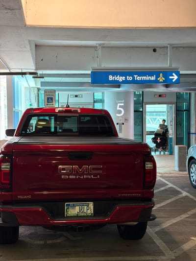 This media file captures a red GMC Denali pickup truck parked in a parking garage. The truck's tailgate displays the "GMC DENALI" badging, and the license plate reads "J&LP S NORTH CAROLINA" with "NC SURVEYORS" and "MAY 22" visible. The word "WILLIAMS" is also present on the tailgate.

The setting is an indoor parking garage, suggested by the concrete structure and overhead lighting. To the left of the truck, a blue sign with an arrow and an airplane symbol points towards "Bridge to Terminal." Further down the garage, a sign indicates "Level 5." A person in a wheelchair is visible through a glass doorway in the background. A sign for "RESERVED PARKING" with the wheelchair accessibility symbol is also present. The lighting suggests it could be daytime, but the enclosed space makes it difficult to determine the exact time of day or weather. The overall scene appears to be a functional space within an airport or similar transportation hub, focusing on the parked vehicle and directional signage.