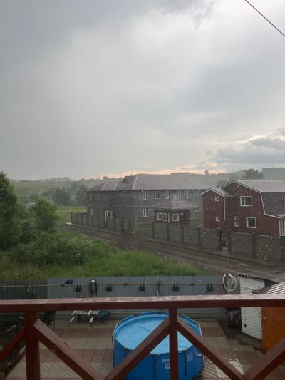 The image captures an outdoor scene in Solontsy, Russia, during what appears to be a light snowfall or heavy rain with visible precipitation in the air. In the foreground, a wooden railing of a balcony or porch frames the view. Beyond the railing, a section of paved patio with a checkered pattern is visible, featuring a bright blue inflatable swimming pool. To the right of the pool, a small white and brown outbuilding is situated.
The middle ground of the image showcases a residential area with two distinct houses. The larger house on the left is a two-story structure with a gray facade and a reddish-brown roof. Adjacent to it is a smaller, red-brick house with a gambrel roof. Both houses have multiple windows. A grey, slat-style fence separates these properties from the street or pathway in front of them. A small, octagonal gazebo with a red roof stands between the two houses.
The background depicts a rural or semi-rural landscape with rolling green hills and trees. Power lines and distant structures can be seen on the horizon, suggesting a developed area further away. The sky is overcast with a predominantly gray hue, indicating cloudy weather. The precipitation is clearly visible as small, white specks scattered throughout the image, more concentrated in the foreground and middle ground.