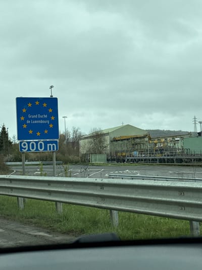The image shows a road sign indicating the border to Luxembourg (Grand Duche de Luxembourg) is 900 meters away. The sign is blue with yellow stars, typical of EU country border signage. A metal guardrail is visible on the right side of the road. Behind the sign, there's a large industrial complex or factory, with tanks and scaffolding. The sky is overcast. The location is likely near Aubange, Belgium, given its proximity to the Luxembourg border.