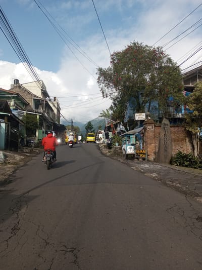 A street view in a residential area. The cracked asphalt road leads uphill. A person on a motorcycle in a red jacket rides away from the viewer. Another motorcycle and a yellow van are farther up the road. Buildings line the sides of the street, some with visible wear. Power lines crisscross overhead. On the right side of the road, a brick wall with a sign reading "the Cibodas" is visible, along with a vendor's cart. A tree with red flowers stands behind the wall. The sky is blue with scattered clouds. Hills are visible in the distance. The location provided is incorrect, as the signs are in Indonesian and the building styles are typical of Indonesian areas.