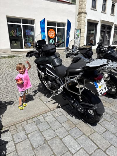 The image captures a sunny day scene in Riedlingen, Germany, as indicated by the motorcycle license plate. A young child, wearing a pink shirt, patterned shorts, and yellow sneakers, stands on a cobblestone street. The child is holding a small, red object in their hand and is looking at the camera with a slight smile, while gesturing with one hand. The backdrop includes a light-colored building with large windows and signage "stille" and "Kultur.Werk.Stadt." Two motorcycles are parked nearby, one of which is prominently featured. The license plate of the motorcycle reads "D AA %7 ST 7," indicating it is registered in Germany. The scene is generally bright and well-lit, highlighting the urban setting and the child's presence.