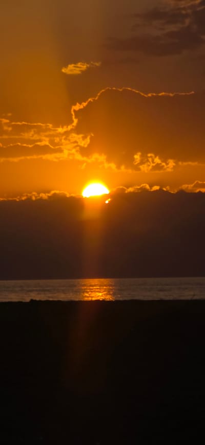 This is a vertical, medium-shot photograph of a sunset over a body of water, possibly a sea or large lake, viewed from a sandy shore. The sun, a bright, glowing orb, is partially obscured by dark, horizontal clouds, with rays of light emanating downwards. The sky above is a gradient of warm oranges and yellows, with smaller, illuminated clouds scattered throughout. The water reflects the vibrant colors of the sunset, creating a shimmering pathway of light. The foreground consists of a dark, silhouetted expanse, likely the shore or beach, with some indistinct texture. There are no people or specific structures visible in the image. The overall scene evokes a peaceful and natural ambiance.