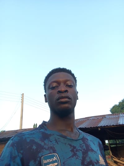 A young Black man is pictured in a selfie taken outdoors under a clear blue sky. He is wearing a dark blue t-shirt with a tie-dye pattern of lighter blue and dark splotches. On the left side of his chest is an Arsenal Football Club logo within a shield. He has short, dark, curly hair and is looking directly at the camera with a neutral expression.
In the background, to the left of the man, is a utility pole with multiple wires extending from it. To the right of the man, and extending across the lower portion of the image, is the roof of a rustic structure, likely a shelter or outbuilding, with corrugated metal roofing that appears aged and rusty. Trees with green foliage are visible behind and to the right of this structure. The overall environment suggests a rural or semi-rural setting. The presence of the utility pole and the structure implies a settled area, possibly within the town of Gindin Dutse, Nigeria, given the context.