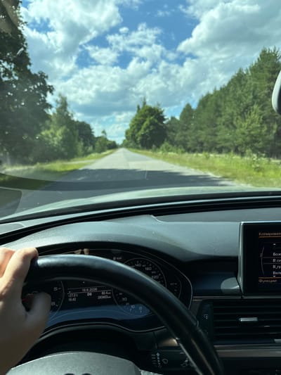 The image is a point-of-view shot from inside a car, looking forward along a paved road that cuts through a wooded area. The sky is blue with fluffy white clouds. The car's dashboard is visible in the foreground, with a hand on the steering wheel. The instrument cluster shows a date of "19.6.2026", a temperature of "+27.5°C", and an odometer reading of "293403 KM". A navigation screen is visible on the right side of the dashboard, displaying text in Cyrillic script. The scene suggests a journey on a sunny day through a rural or suburban environment, likely in Ukraine given the Cyrillic text. There is no indication of the city of Kostiantynivka, Ukraine in the image.