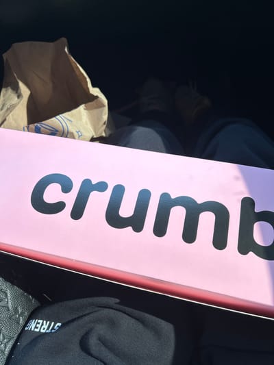 In Aventura, Florida, USA, an overhead view captures a pink box with the word "crumb" emblazoned in large, black, block letters. To the left of the box, a crumpled brown paper bag is visible, with blue designs on its interior. The scene appears to be inside a vehicle, as evidenced by the dark interior and the presence of what looks like a person's legs and feet wearing tan shoes and dark pants in the background. The bottom of the image shows a black fabric with white lettering that reads "STRENG" backwards.