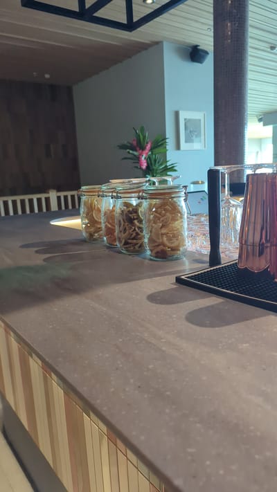 The image shows a close-up view of a bar counter in what appears to be a restaurant or bar in Larkollen, Norway. The counter is made of a speckled grey material. On the counter, there are three glass jars filled with what look like dried citrus slices. Behind the jars, there are stacks of clear glasses and a copper cocktail shaker. A black mat with a textured surface is also visible on the counter. In the background, there is a wall with wooden paneling, a pillar covered in mosaic tiles, and a framed picture on the wall. A plant with pink flowers is also present in the background. The lighting suggests an indoor environment, possibly during daylight hours. There are no people visible in the image.