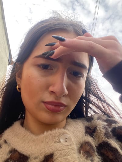 The image displays a close-up, low-angle portrait of an adult female, reportedly located in Broxbourne, United Kingdom. She has long, dark brown hair, dark eyes, and full lips with reddish-brown lip color. Her facial expression is neutral, with a slight furrow in her brow. She is wearing a fluffy cardigan or jacket featuring a beige and brown leopard print pattern, with one white button visible on the front. A gold-colored hoop earring is present in her left ear. Her right hand is raised, with four fingers lightly touching her forehead above her left eyebrow. The fingernails on this hand are long, pointed, and painted dark grey or black. The background consists of an overcast sky with visible utility wires in the upper right, and a light-colored building facade partially visible on the far left.