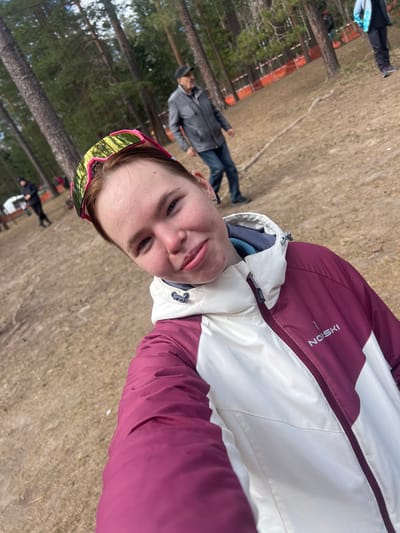A selfie photograph taken outdoors in Lesnoye, Russia, captures a person wearing a white and burgundy ski jacket and sunglasses with a pink frame. The individual is positioned in the foreground.

In the blurred background, trees are visible on the left, and two people are seen. One is wearing a grey jacket and dark pants, while the other is on the ground near a white object. Beyond them, a portion of a fence or structure painted in red and orange can be identified. The ground appears to be covered in dry grass.