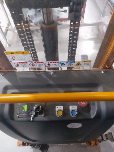 The image displays a detailed, slightly angled overhead view of the control panel and a portion of the mast mechanism of an industrial electric stacker, manufactured by Soosung Industrial Co., Ltd. The scene is located indoors, likely within a manufacturing or warehousing facility belonging to Soosung, or a facility utilizing their equipment, in Gimpo-si, Gyeonggi-do, South Korea.

The upper section of the image shows the machine's lifting mast, featuring a central dark hydraulic cylinder and two vertical lifting chains. A clear polycarbonate protective shield is positioned in front of these components. Through the shield, a blurry background reveals an indoor environment with light-colored flooring and what appear to be stacked white industrial sacks or packaging.

Below the clear shield, a series of Korean safety warning decals are affixed. These include pictograms and text indicating "Caution," "Pinch Point Hazard" (for hands, feet, and head), "Falling Object Hazard," "No Riding," and "Operation Prohibited for Unauthorized Personnel" or "Complete Safety Training."

The lower portion of the image is dominated by the machine's dark gray control panel, topped by a horizontal yellow handrail or support bar. On the control panel, from left to right, are:
- A rectangular display showing a green battery charge indicator and the website "www.soosung.co.kr".
- An ignition switch with a key inserted.
- A yellow circular push-button labeled with an upward arrow and "↑ UP".
- A blue circular push-button labeled with a downward arrow and "↓ DOWN".
- A prominent red circular emergency stop button.
- A white oval sticker with Korean text, likely containing model or certification information.

No personnel are visible in the frame, and the machine appears to be in an idle state.