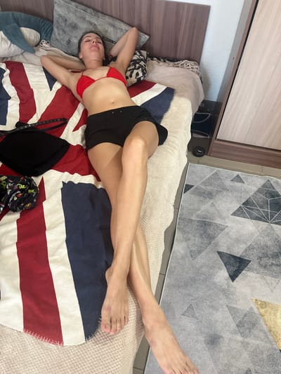 A young woman is lying on a bed with her arms behind her head and her legs crossed. She is wearing a red bikini top and black shorts. The bed is covered with a Union Jack-themed blanket, and there are pillows and a rolled-up pair of patterned shorts on the bed. A black bag and a belt are also on the bed. The scene appears to be indoors, likely in a bedroom, with a wardrobe visible in the background. The floor is tiled and covered with a geometric patterned rug. The overall impression is one of relaxation.