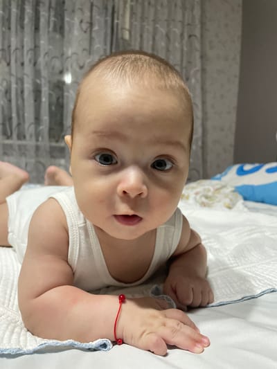A close-up, eye-level shot shows a baby lying on its stomach on a white, textured surface, likely a bed. The baby is wearing a white undershirt. Its face is turned towards the camera, with large, dark eyes and a slightly open mouth. A thin red string bracelet with two small red beads is visible on its left wrist. In the background, sheer curtains with a patterned design are visible, and a blue and white pillow lies to the right of the baby. The lighting is soft, suggesting an indoor setting. There is no indication of the specific location in Fălești, Moldova.