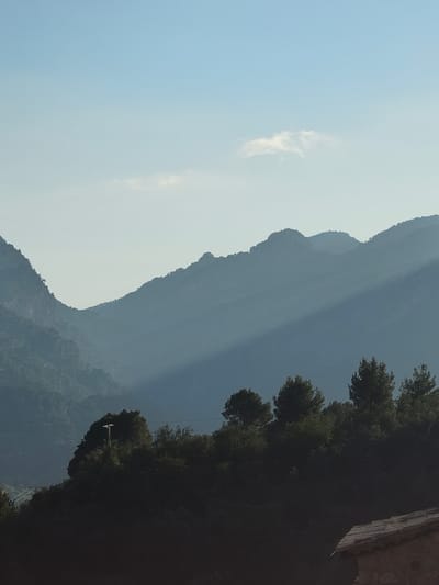 The image captures a panoramic view of a mountainous landscape in Fornalutx, Spain, under a clear, pale blue sky. A single, wispy white cloud is present in the upper right sky. Multiple layers of mountains, heavily forested, recede into the distance, appearing progressively lighter and bluer due to atmospheric perspective. A distinct diagonal beam of sunlight illuminates a section of the mid-ground mountains, creating a bright band across the otherwise shadowed or hazy terrain. In the dark foreground, a ridge densely covered with evergreen trees and foliage is visible. On this ridge, a utility pole with a light fixture stands on the left. The lower right corner of the frame reveals the tiled roof and stone wall of a building. No people or animals are visible.