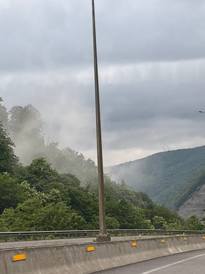 A low-angle, eye-level, full shot captures a view of a highway curving through a lush, green, mountainous landscape on a heavily overcast day. The foreground is dominated by a concrete barrier with several reflective yellow markers. A tall, slender light pole stands vertically in the middle of the frame, extending towards the cloudy sky. Beyond the highway barrier, a dense forest covers the slopes of rolling hills and mountains. Wisps of fog or mist are visible amongst the trees, creating a hazy atmosphere. The sky is filled with dark, heavy clouds, obscuring any direct sunlight. The image appears to be taken from a moving vehicle on the highway, suggested by the perspective and the slight blur in the background. There are no people visible in the scene. The location is identified as Tsakva, Georgia.
