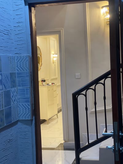 The image, rotated to its correct upright orientation, depicts an interior view within a building, possibly a residence or commercial space. In the immediate foreground, a dark wooden beam or railing horizontally frames the scene. Below this beam, a wall features a textured, light blue-grey plaster finish, partially covered by decorative tiles arranged in a grid pattern. These tiles display various geometric designs, including chevrons, stripes, and abstract shapes, in shades of blue, white, and grey.
Beyond the foreground railing, a black wrought iron balustrade with ornate scrollwork extends upwards, suggesting a staircase or an upper-level landing. The surrounding walls are painted a light color, likely off-white, and feature classic decorative moldings. A white electrical switch is mounted on one of these walls.
Further back in the scene, a modern wall sconce with a gold finish casts light on an upper wall. Below it, a white doorway with trim leads into an adjacent room. Inside this room, another similar wall sconce is visible, alongside a golden-framed oval mirror mounted on the wall. A white rectangular unit, possibly a refrigerator, cabinet, or an electrical power box with visible wiring, stands within this room. The floor of the lower level, visible towards the right, is covered with glossy, marble-patterned tiles, leading to what appears to be the bottom of a staircase.
No people are present in the image, and no specific activity is occurring. The lighting suggests an indoor setting illuminated by artificial sources, possibly indicating evening or a reliance on interior lighting. No discernible text is visible anywhere in the image. The architectural elements appear modern and well-maintained.